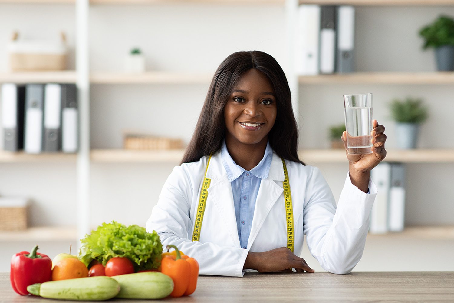 Young female african american nutritionist holding glass of water while sitting at table with fruits and vegetables. Crystal Seals is a Registered Dietitian qualified to work with medical conditions like Diabetes, cardiovascular (heart) disease such as high blood pressure, high cholesterol, and arteriosclerosis. 